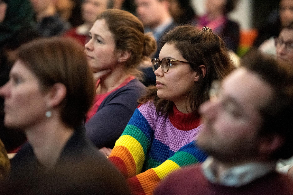 Jonge vrouw in regenboogtrui tussen collega's tijdens bijeenkomst. Maak kennis met de mensen van Buitenlandse Zaken