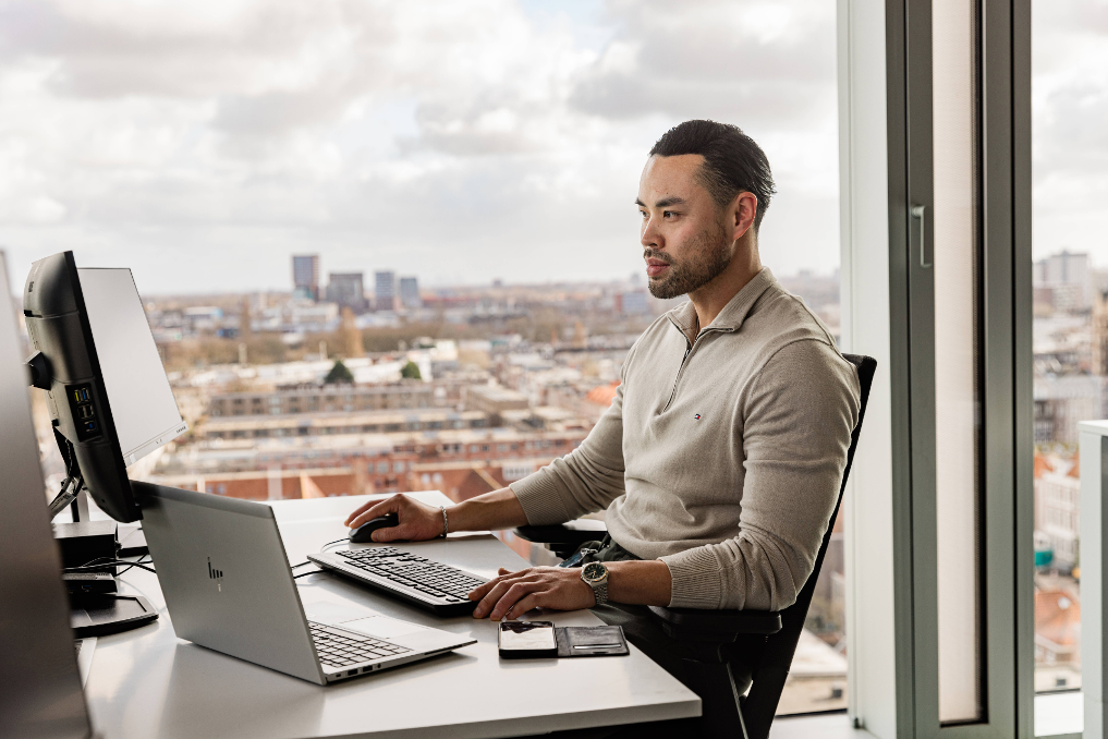 Een collega bij het ministerie van IenW werkt aan de laptop. Achter hem de skyline van Den Haag