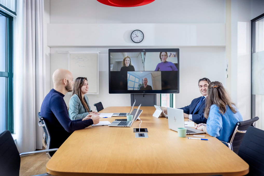 Roos en collega's in overleg, met mensen in de zaal en mensen op het scherm. Ze zijn jurist bij de Rijksoverheid